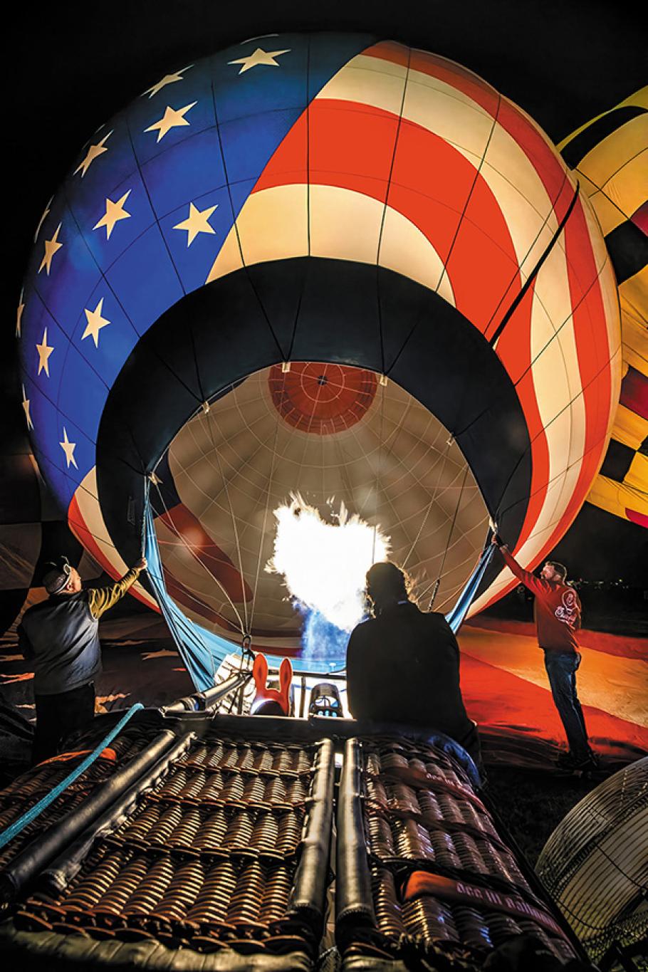Below a dark sky, three people attend to the inflation of a balloon envelope attached to a wicker basket. Both the envelope and basket lie on their sides on the ground. The envelope bears the red, white, and blue stars and stripes of the American flag and is inflated.