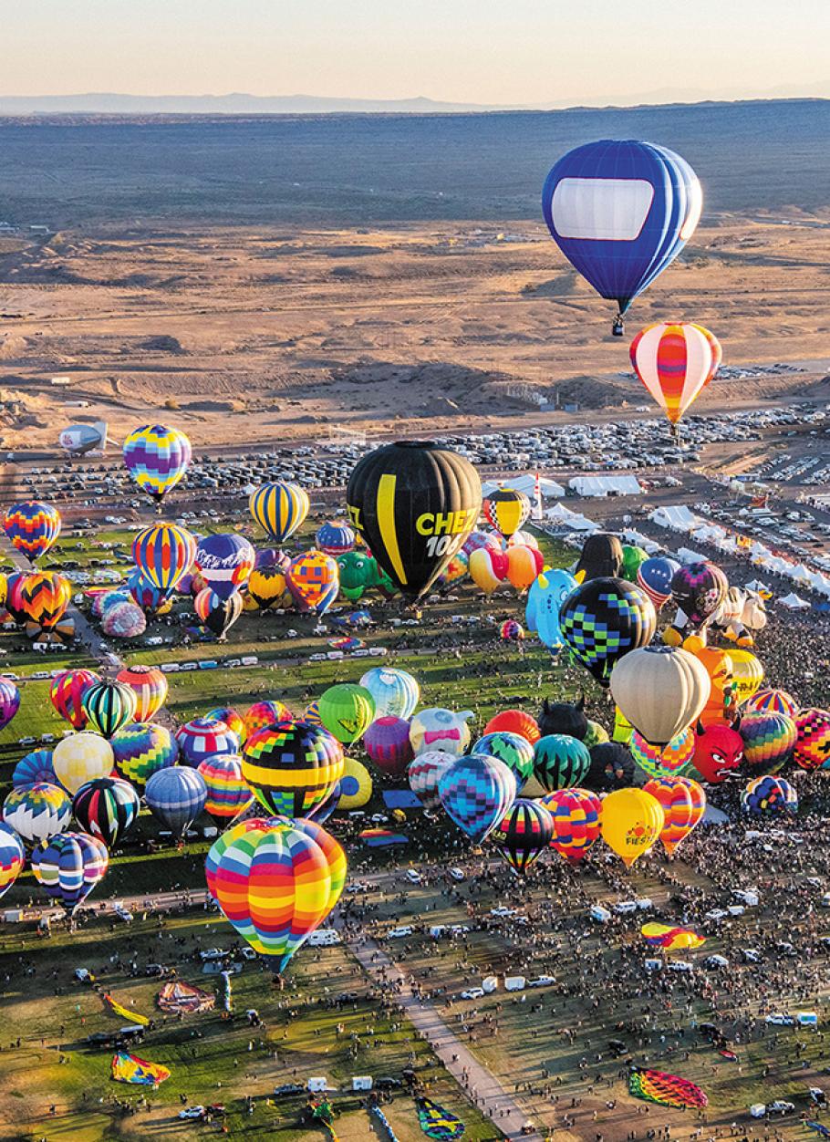 Dozens of multi-colored hot air balloons ascend into the sky--staggered at different altitudes against the backdrop of the desert below and the festival grounds crowded with people.