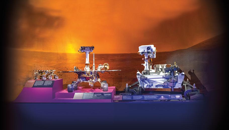 Three robotic Mars rovers are displayed on a purple shelf with a bright red photo of Mars landscape in the bakcground. The first on the far left is the smallest, with six wheels and a simple chassis covered in solar panels. The second, middle rover is slightly larger, with instruments mounted on a short pole resembling a "head." The third, largest rover, which is the size of a small car, has complex intruments covering its large chassis as well as a pole resembling a head.