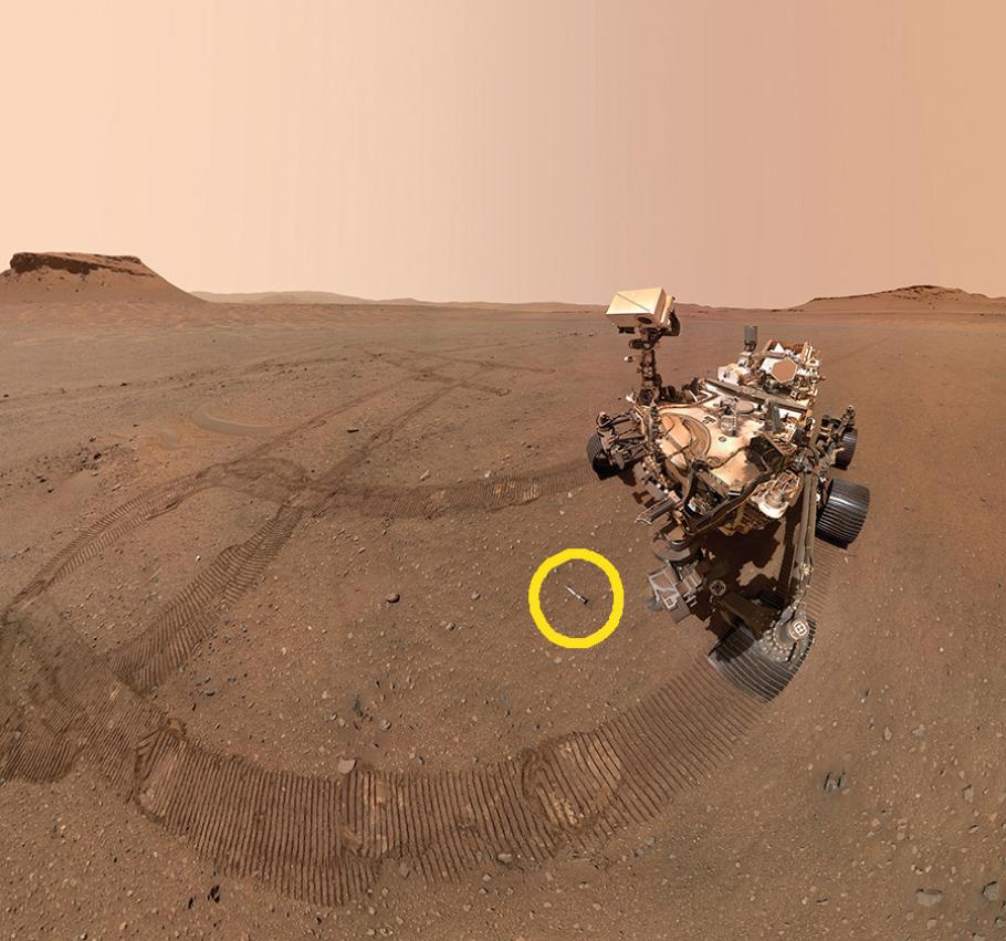 A large robotic rover leaves tracks in the red Martian desert, under a red-tinted sky. The rover has a large rectangular body supported by several wheels, emrging from the body is a camera on a pole, giving it a neck and head look. In this image, the camera is pointed to the ground where a small tube is on the ground behind the rover. The tube is highlighted by a superimposed yellow circle. 