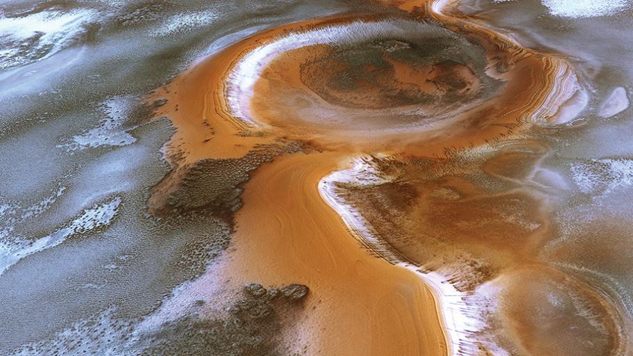 A large round crater with craggy edges has a dark brown interior, with some white frost visible at the bottom. Around it the dirt patterns of Mars swirl, like water on sand when the tide goes out.
