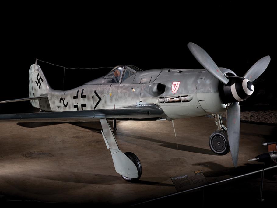 A German fighter aircraft is painted dull grey, with some black markings including a Nazi swastika. A large gun in mounted near the nose of the plane, which has a single large propeller.