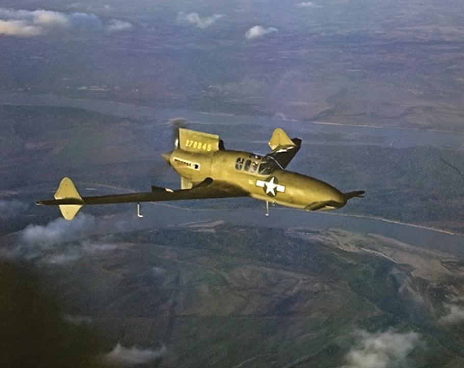 A dark green aircraft flies above a green landscape. The airplane features two short wings on the nose, two large swept-back wings near the rear, with a propeller mounted behind the pilot's cockpit.