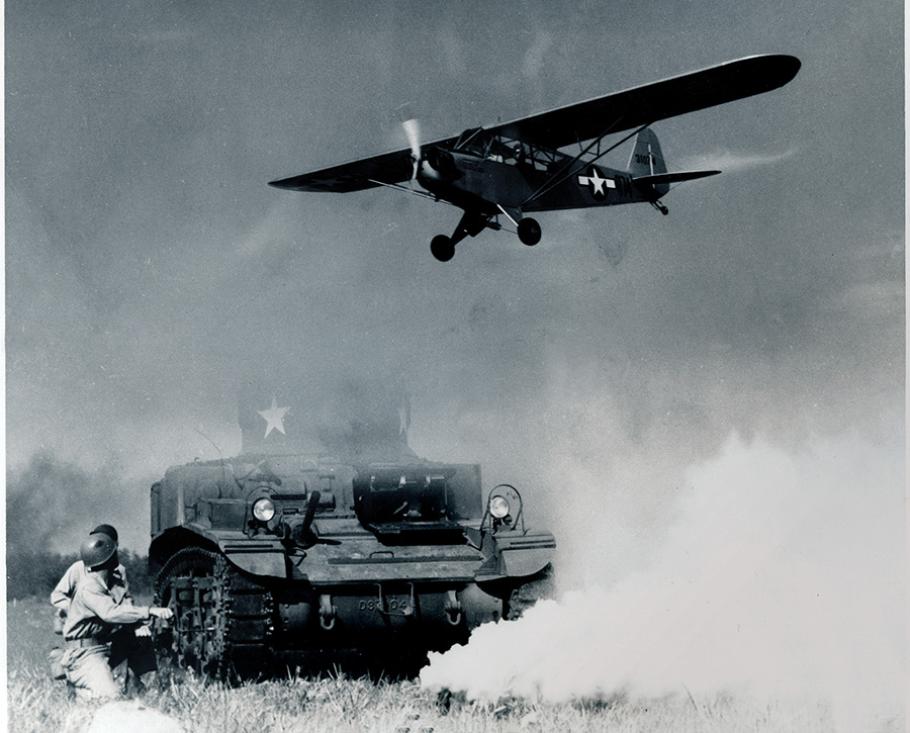 Black-and-white photo of a small, unarmed single-propeller aircraft flying over two U.S. soldiers crouched behind a tank.