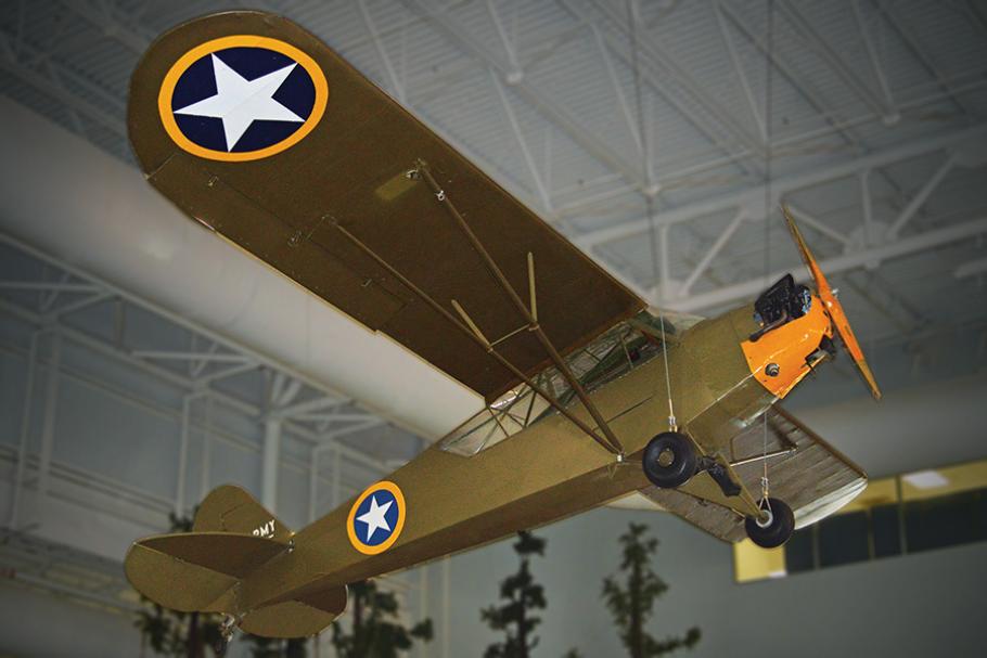 A small, unarmed military airplane is suspended from the ceiling of a museum. The aircraft is painted dark brown with an orange nose and U.S. Army stars on its chassis and wings.