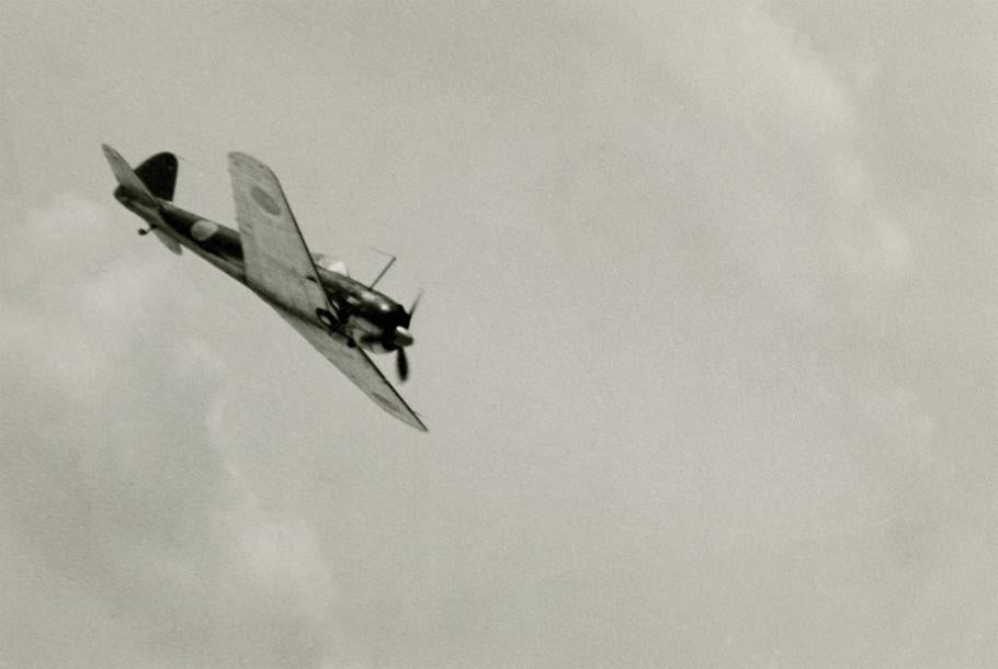 A black-and-white photo of a single-propeller fighter aircraft, with Japanese markings visible on its wings. The airplane is flying in an empty sky, at a downward angle.