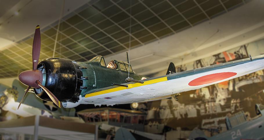 A single-propeller fighter aircraft is suspended by wire from the ceiling of a museum. The airplane has a dark green fuselage, but the underside of the wing is white, with a bright red "rising sun" marking of the Japanese air force.