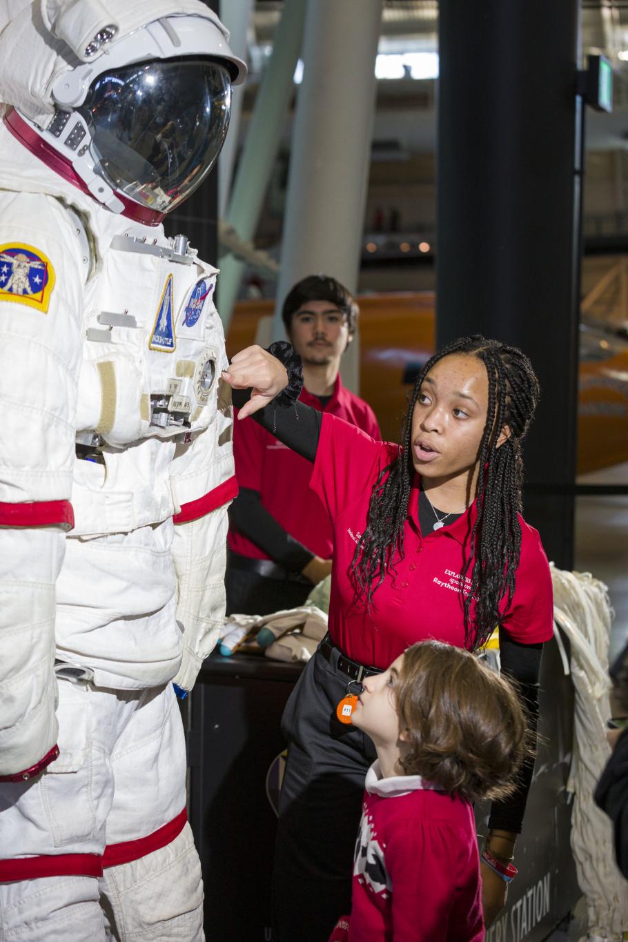 A museum staff points to different components of a space suit while young visitors look on.