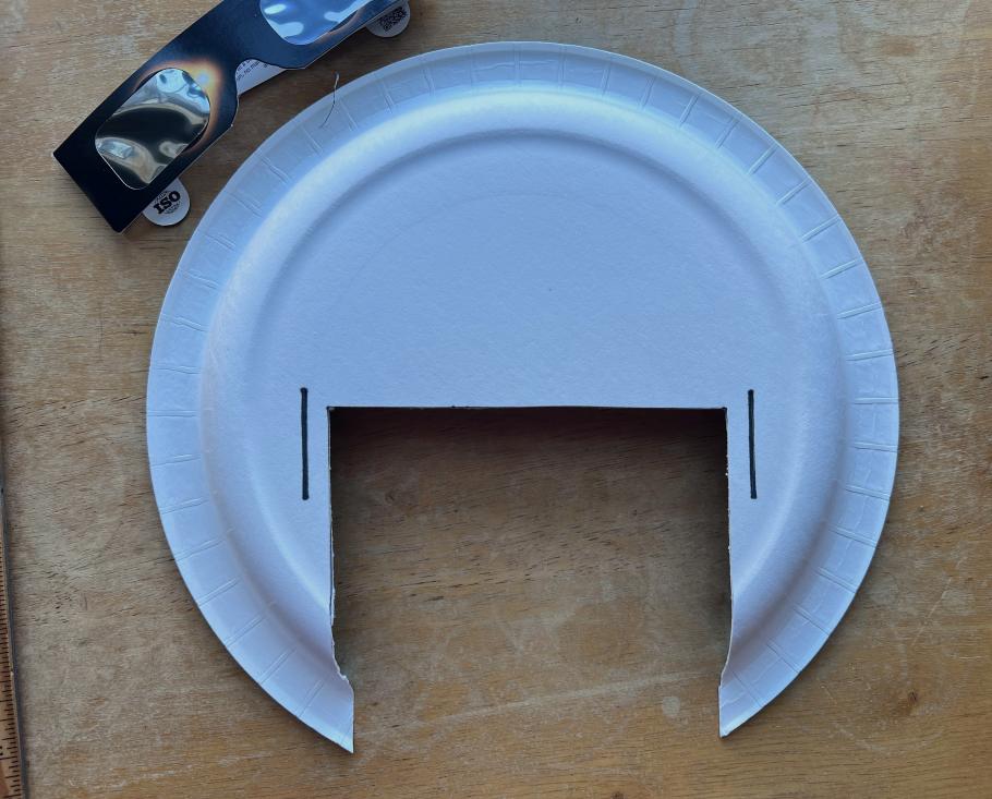 Photo of white paper plate with a section cut out of it. There are a pair of solar eclipse glasses at the top of the photo that you can see partially. 