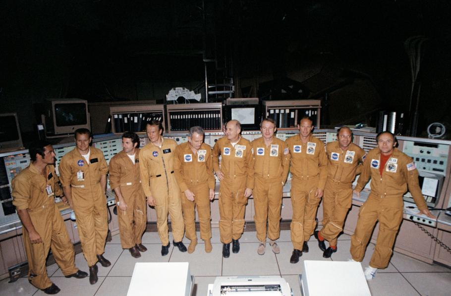 10 middle-aged men wearing yellow-brown jumpsuits stand in Mission Control. Some are looking at the camera and others are looking away.