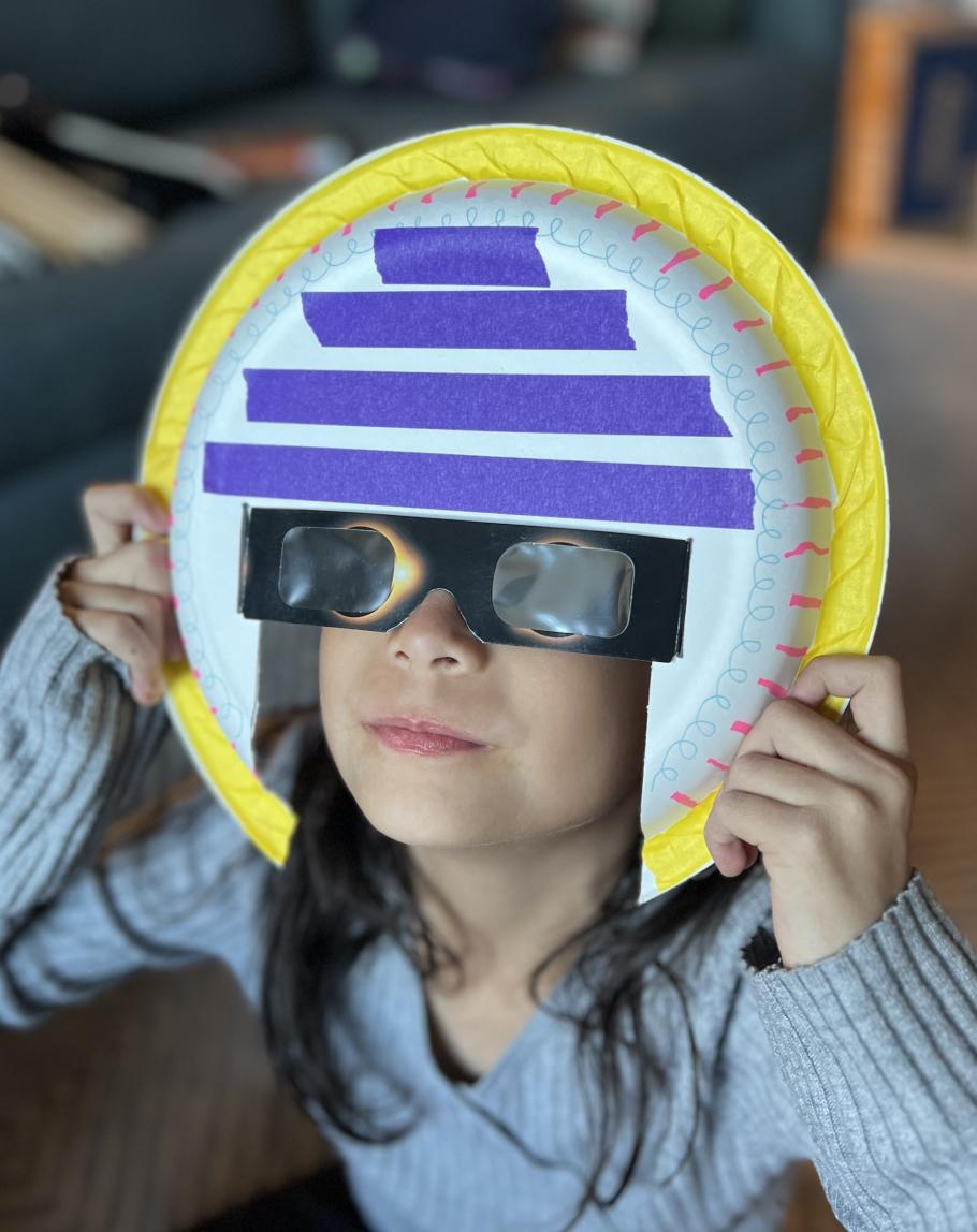 Photo of young girl holding up to her face a paper plate cut to hold solar eclipse glasses - it is an eclipse mask. She has the glasses held up to her eyes and is looking through the glasses. The  eclipse mask is decorated with yellow and purple craft tape, and with blue and pink markings.