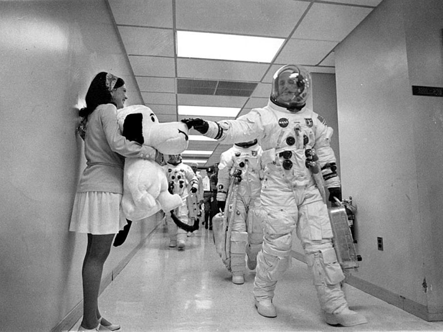Black and white image of a astronaut walking down a hallway. Other astronauts are walking behind him. A woman stands next to him holding a large teddy bear.
