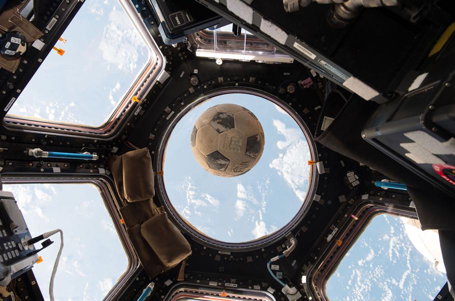 A soccer ball floats inside the International Space Station surrounded by a panoramic array of windows that provide a view of Earth and space.