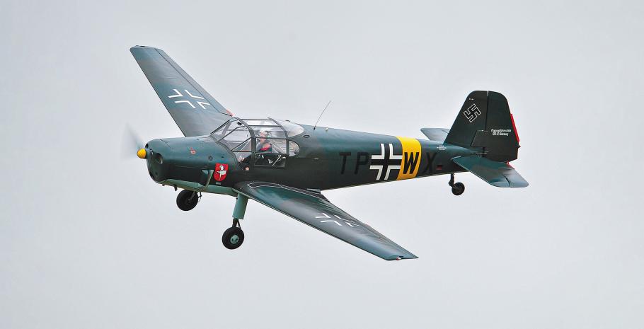 A small aircraft painted dull green with German WW II markings flies in the air at a modern-day airshow.