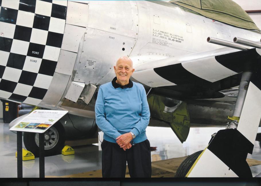 An elderly man wearing a light blue sweater poses next to a P-47 aircraft on display at a museum.