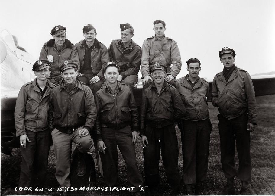 A group of American airman pose for a casual photo during World War II. They stand in two lines, leaning on each other suggesting commradarie. Many wear leather jackets and military caps, though they's tipped to one side. 