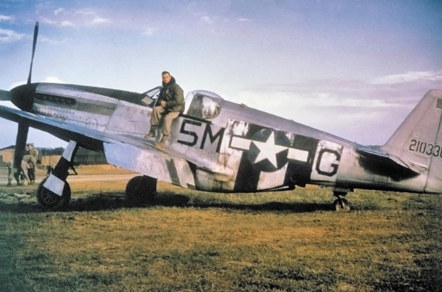 An American pilot sits on the fuselage of a World War II fighter aircraft, with a metallic gray fuselage.