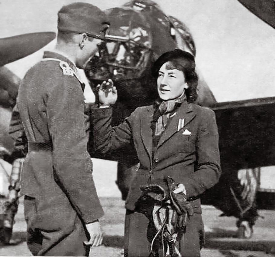 A  woman in her 40s speaks with a German soldier during World War II, a bomber aircraft parked behind them. She is looking up at the soldier, gesturing and his body language is one of respect, intent on what she's saying.