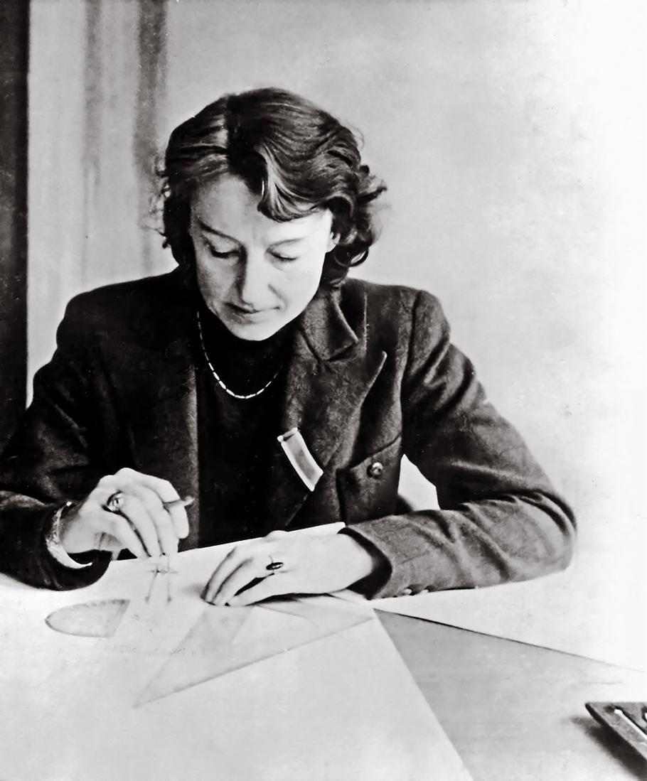 A woman in her 40s sits working at a drafting table. The photo is in black and white, and the woman wears a tweed blazer, pearls, and slightly styled hair, suggested it was taken in the mid-20th century.