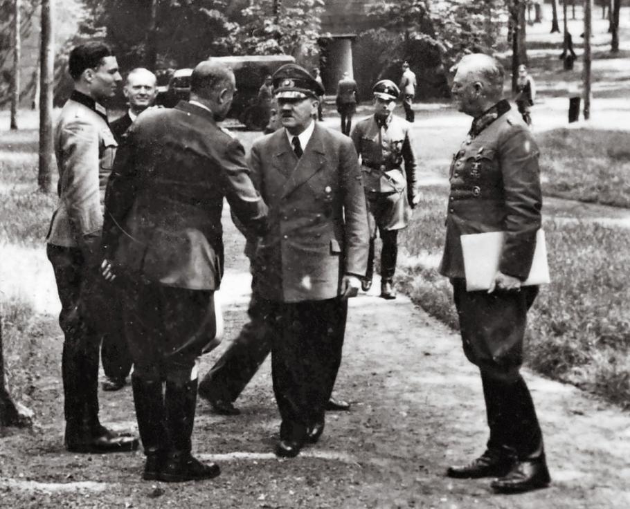 A group of high-ranking German military officers speak with Hitler. The photo is taken from afar in a wooded area. The men are wearing military uniforms. Hitler stands stiffy, shaking another man's hand. His signature mustache is evident in the black and white photo. 