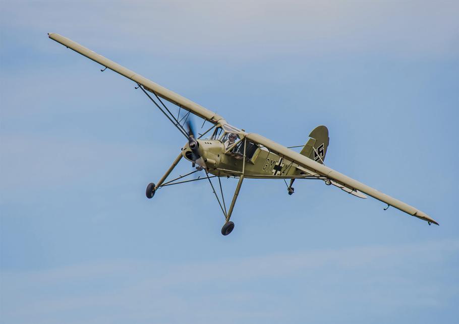 A restored small German liasion aircraft painted with World War II markings flies in the sky over a modern-day airshow. It is a slender olive plane with a propellor ont he nose. 