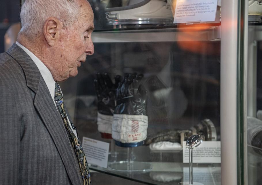 An elderly individual, Bill Anders, looks at a watch on display in a case amongst other space artifacts including gloves and helmets.