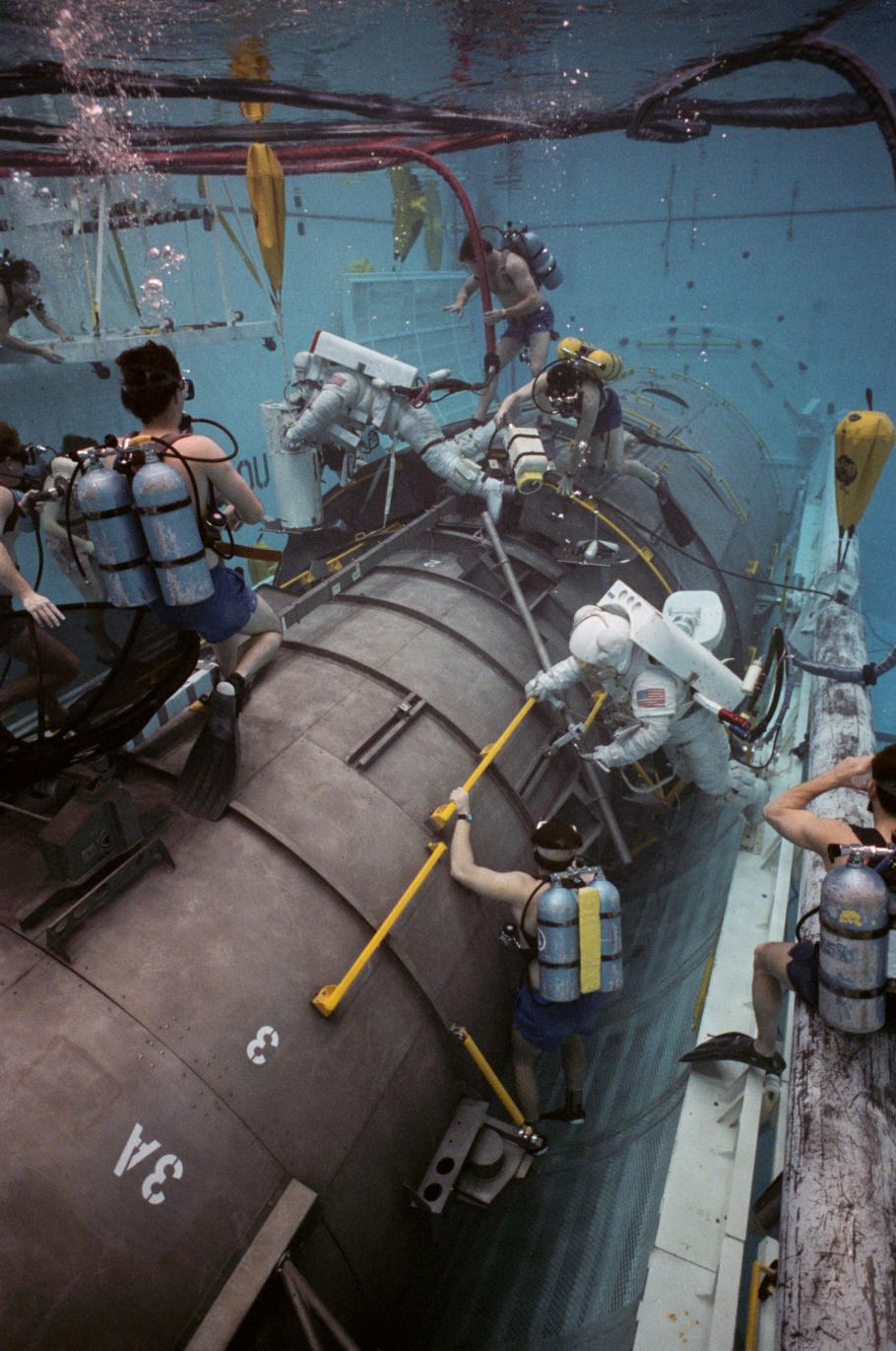 Astronauts participate in underwater training at NASA's Neutral Buoyancy Lab, practicing skills on a life-sized replica of a spacecraft module. Divers, wearing scuba gear, assist and observe the simulation.