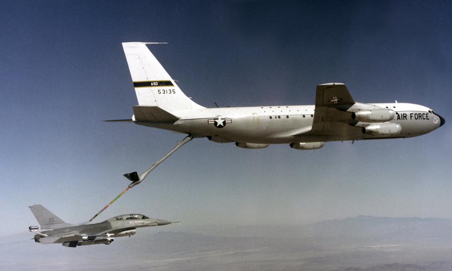 A gray, single-engine military jet is docked to the refueling probe of a much larger, four-engine jet airplane flying above it--during the day. The larger airplane is marked with the words "Air Force" and the USAF stars-and-bars insignia.