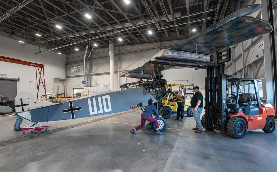 Inside a large aviation hangar with a polished concrete floor, three people work to attach the wing of an early airplane to the green-painted fabric and wood fuselage. A hydraulic lift holds with wing securely as it's lowered toward the fuselage.