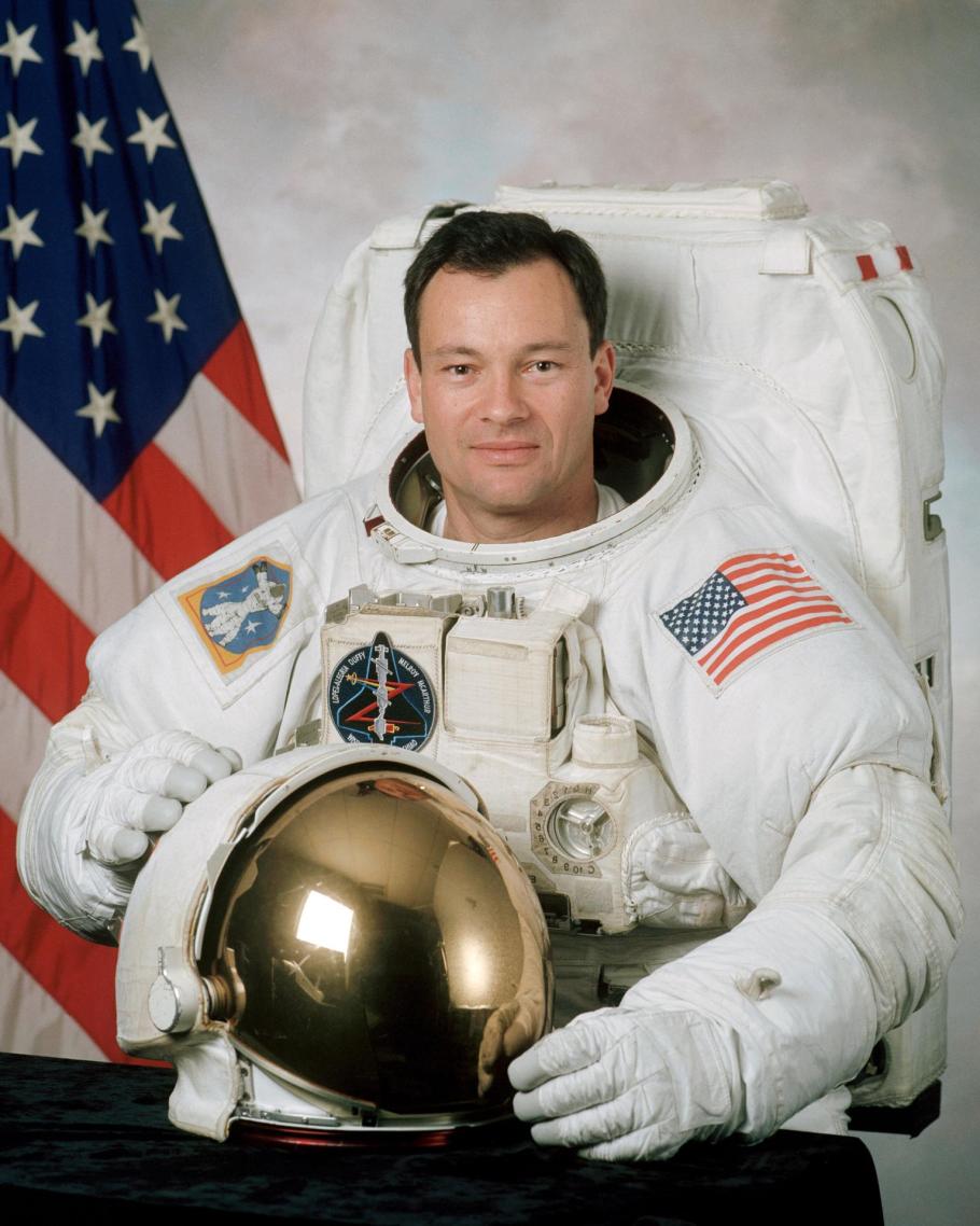 A portrait of astronaut Michael López-Alegría in a white space suit, holding his helmet under his left arm. The suit features various patches, including a NASA emblem, an American flag, and mission insignia. An American flag is visible in the background.