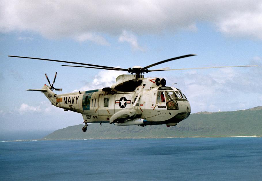 A white and gray helicopter flies over water with the coastline in the background.