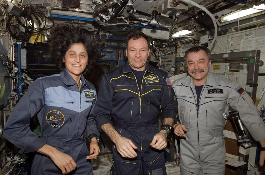 Three astronauts wearing space agency uniforms with mission patches are inside a spacecraft. The background is filled with various equipment and instruments indicative of a space station’s interior, such as control panels, wires, and storage compartments.