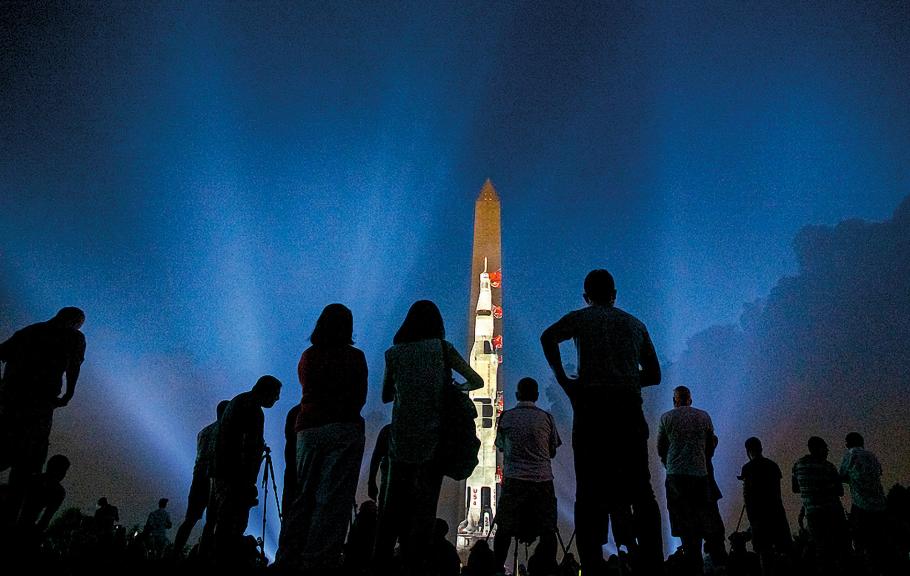 An image of a white, multi-stage rocket is projected onto one side of the Washington Monument at night. Ground lights illuminating the Monument color the sky a midnight blue. The dark silhouettes of several people standing in front of the Monument are visible in the foreground.