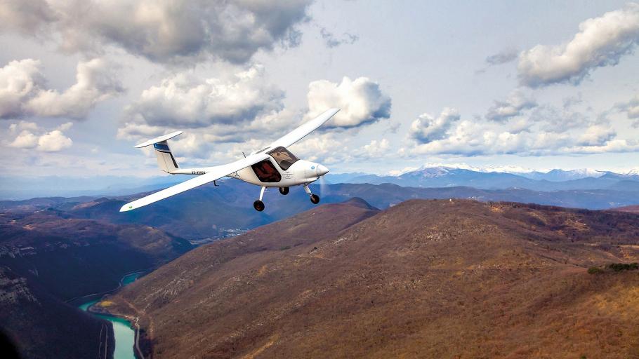 A small, white airplane flies low over a river and brown desert hills. Large white clouds are above the airplane. The airplane is a high-wing monoplane with a single propeller-driven engine and tricycle landing gear.