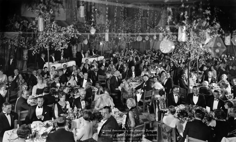 A black and white photography from the 1920s showing a banquet room full of well dressed, middle aged, white people. The men where tuxes, the women pearls and cocktail dresses. The room itself is lavish, with streamers tumbling from the ceiling and trees on the floor. 