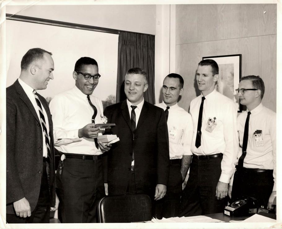 Group of six people in formal attire smiling and interacting in an office setting, with one individual presenting a small box to another.