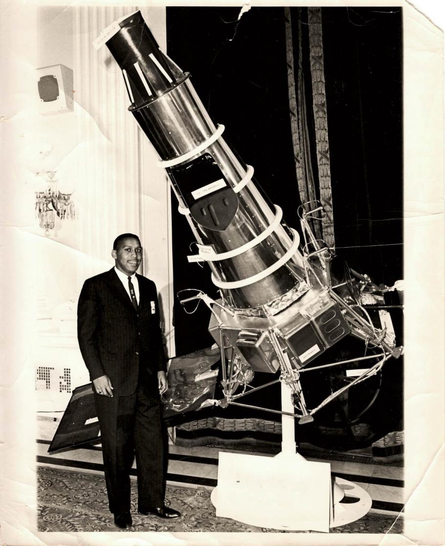 Photograph of an individual standing next to a small satellite spacecraft on display.