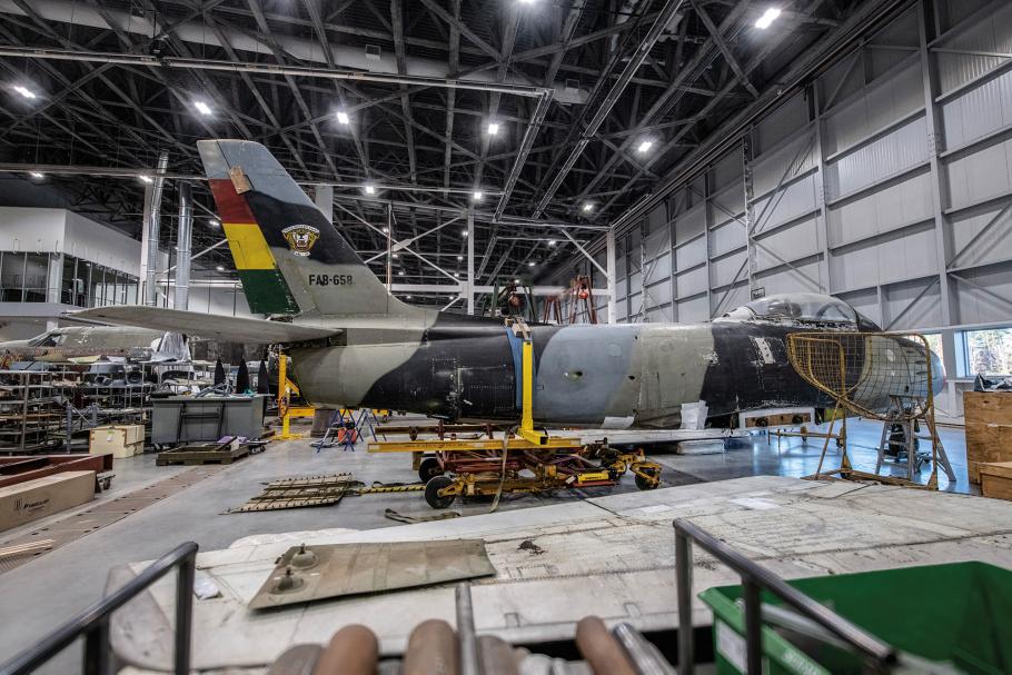 An F-86 military airplane is parked inside a cavernous restoration hangar. The airplane is intact, but its wing has been removed.