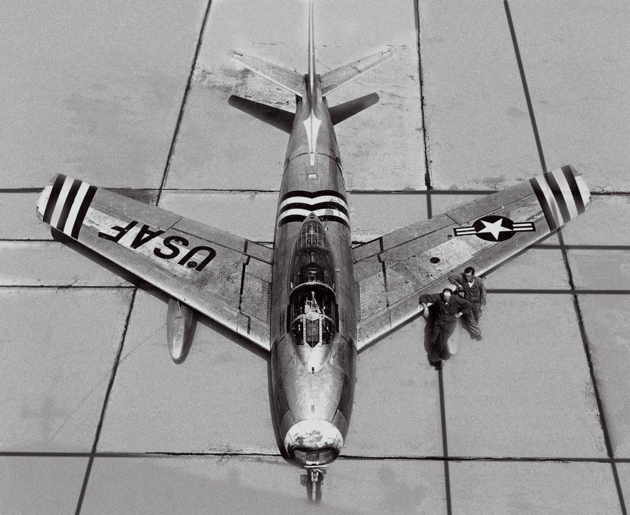 A black-and-white image of a single-engine, jet-powered airplane parked on an airport ramp during the day. The silvery metal skin of the jet is covered in military markings: black and white stripes and the letters: USAF.