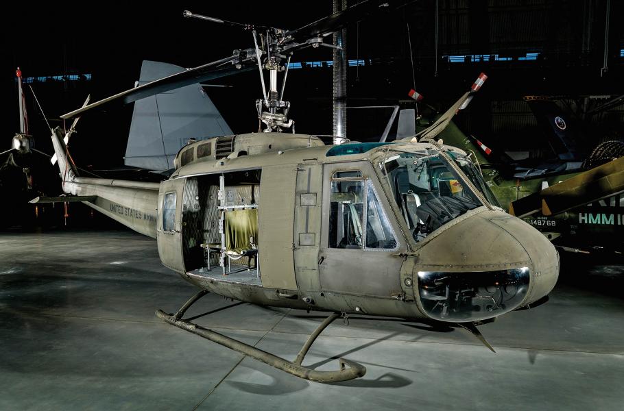 A helicopter painted in an olive green color sits on the floor of a large museum display hangar. Stenciled letters on the helicopter's tail spell out United States Army.