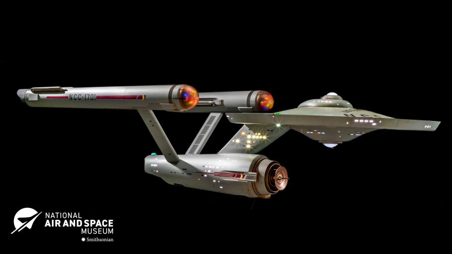 A studio model of the spaceship Enterprise from Star Trek floats against a black background. The ship as a disk shaped center with three horizontal cylinders trailing behind.  