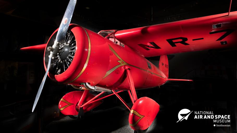 A red monoplane with a propeller sits at a jaunty angle against a black background. 