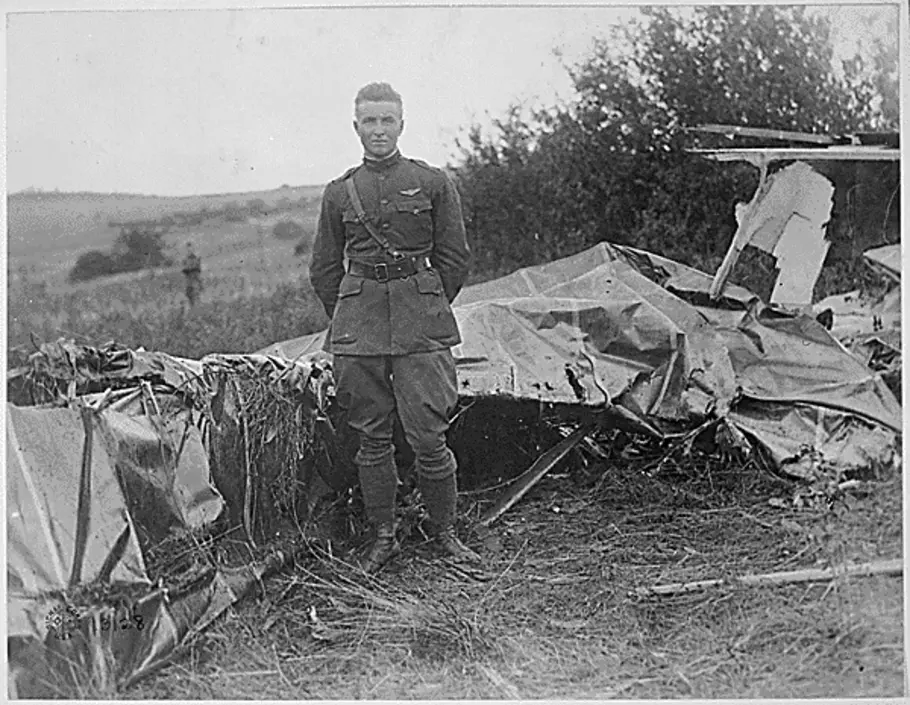 A young white man in a World War I military uniform, stands in front of a wreckage of a balloon. 