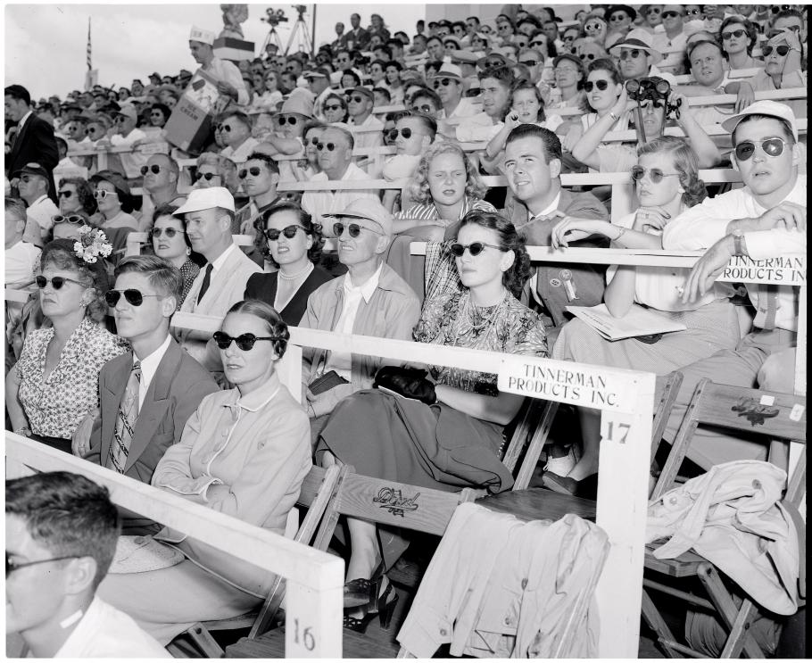 Spectators in a grandstand look to their left. Many wear sunglasses.
