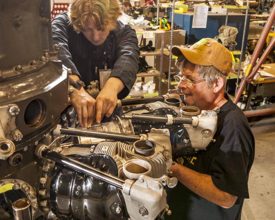 Two white, male Museum specialists work together to perform restoration work on an engine of a World War II bomber aircraft. One of the men is holding his hands below the visible section of the engine while the other man has his fingers above the visible engine section.