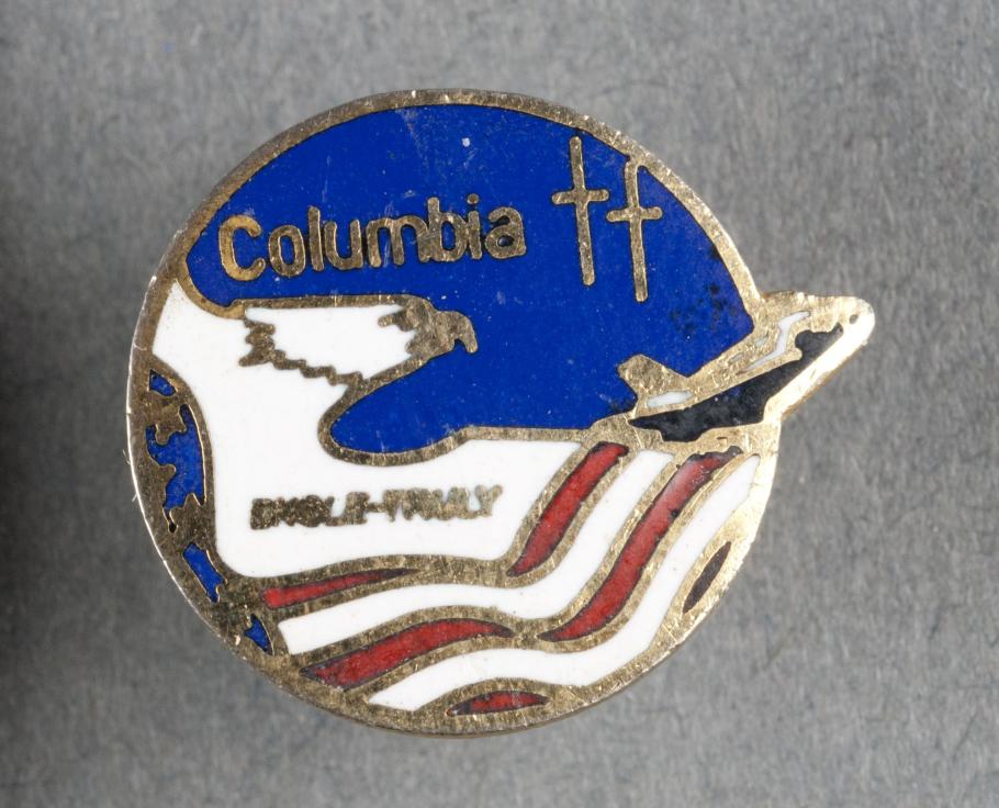 A small gold pin decorate with red, white, and blue. It depicts an artistic interpretation of an eagle flying across an American flag and a space shuttle flying in space.