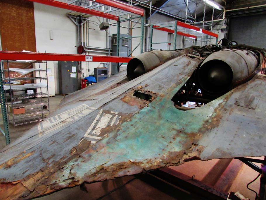 Aft, diagonal view of a gray World War II-era aircraft which reveals green paint under where a fairing section was located prior to its treatment.