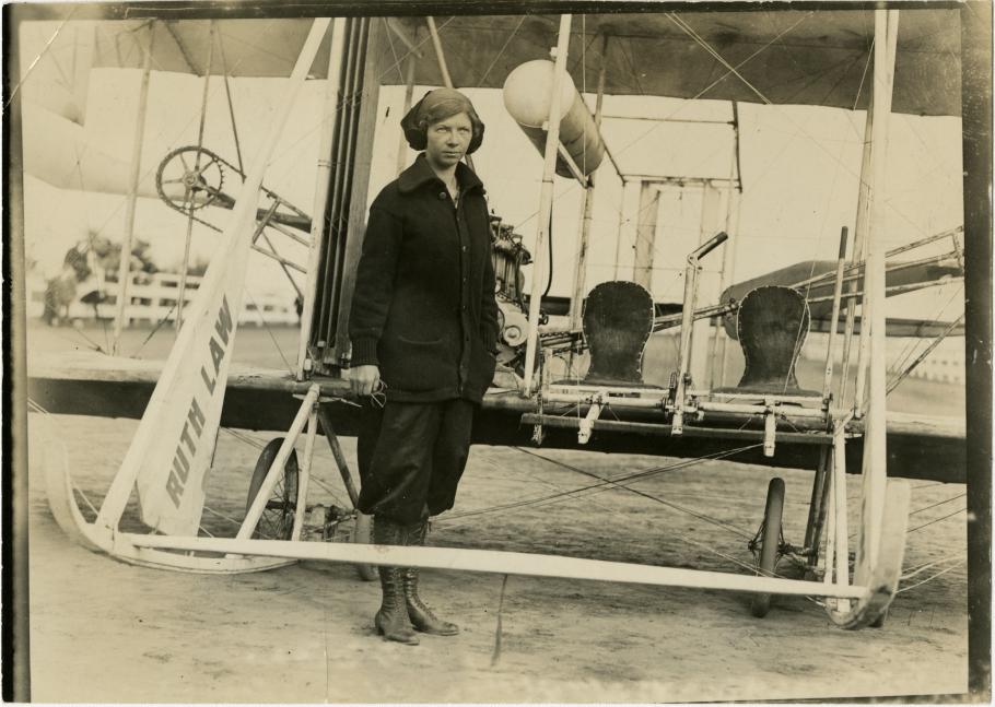 Woman in Dark Buttondown Sweater and Laceup Boots stands in front of early aircraft, with "Ruth Law" written on a part of the aircraft