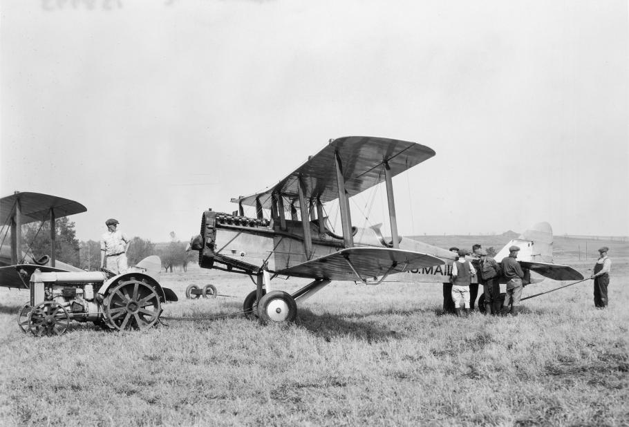 Side view of a light-colored, metal biplane behind a tractor in a field.