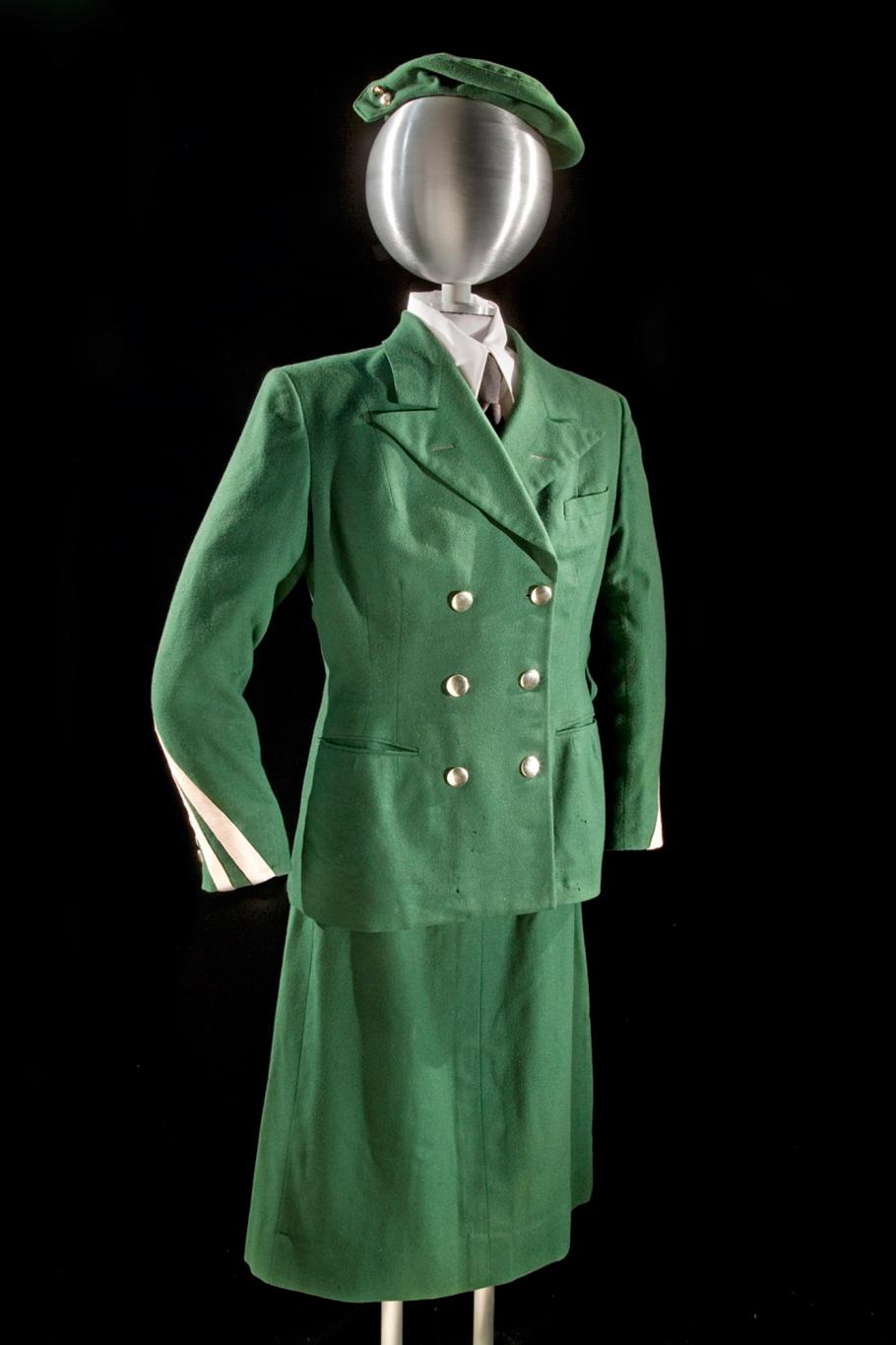Front view of green stewardess uniform with green jacket with three pockets and six brass buttons, a green stewardess cap, and green skirt. White scarf worn on neck. Styled on mannequin.